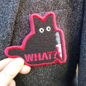 Peut inclure: Écusson noir en forme de chat avec un contour rouge et le mot "WHAT?" en rouge. Le chat tient un couteau. L'écusson est sur un fond en tissu gris.