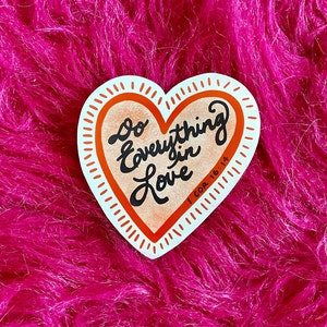 Puede incluir: Una pegatina en forma de corazón con fondo melocotón y contorno blanco. La pegatina dice "Haz todo con amor" con una referencia bíblica, "1 Cor 16:14".