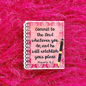 May include: A pink and white notebook-shaped sticker with the text "Commit to the Lord whatever you do, and he will establish your plans. Proverbs 16:3." A black and white polka-dotted pen is on the right side of the sticker. The background is a bright pink, fuzzy material.