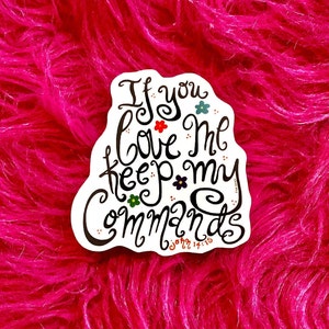 May include: White sticker with black lettering that reads "If you love me keep my commands John 14:15". The sticker has small flower accents and is on a pink fuzzy surface.