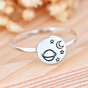 May include: A silver ring with a round design featuring a planet, crescent moon, and stars.