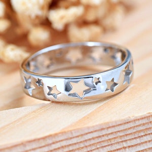May include: A silver ring with star cutouts. The ring has a polished finish and a band with multiple star-shaped openings. The ring is on a wooden surface. The text "PerfectEngagementUS" is at the bottom of the image.
