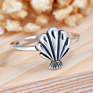 May include: A silver ring with a detailed seashell design. The ring is on a wooden surface.
