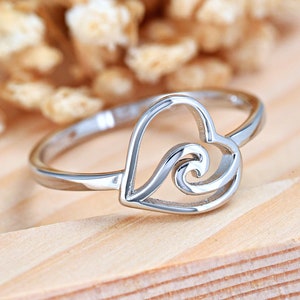 May include: Silver ring with a heart-shaped design featuring a wave inside. The ring is on a wooden surface.