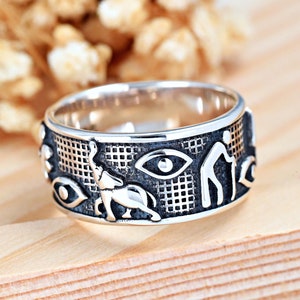 Puede incluir: Un anillo de plata con un diseño intrincado que presenta ojos, una persona y un animal. El anillo es ancho y tiene una superficie texturizada.