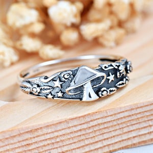 May include: A silver ring with a mushroom design and stars and leaves. The ring is on a wooden surface.
