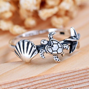 May include: Silver ring with a sea turtle, seashell, and starfish design. The ring is on a wooden surface.