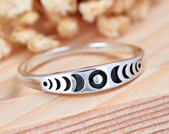 May include: A silver-toned ring featuring a series of engraved moon phases along the outer band. The design displays a full moon in the center, flanked by waxing and waning crescent shapes, with a polished metallic finish and dark recessed details.