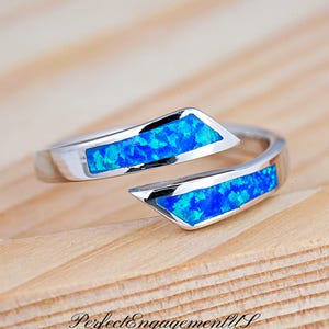 May include: A silver-colored ring with two sections of bright blue opal inlay. The ring has an open design, with the opal sections set in a geometric shape. The ring is displayed on a light wood surface.