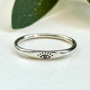 May include: A silver ring with a simple eye design. The ring is smooth and has a slightly rounded shape.