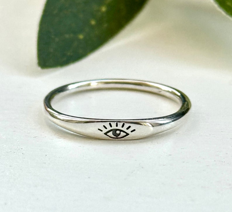 May include: A silver ring with a simple eye design. The ring is smooth and has a slightly rounded shape.