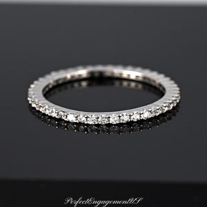 May include: A silver-toned eternity band ring featuring a continuous row of small, clear, round-cut stones set along the entire circumference of the metal band. The text PerfectEngagementUS is visible at the bottom of the image.