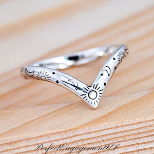 May include: A silver-colored V-shaped ring with celestial designs. The ring features sun, moon, and star motifs. The ring is on a wooden surface.