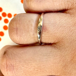 May include: A thin silver band ring with an eye design engraved on the top. The ring is worn on a finger.