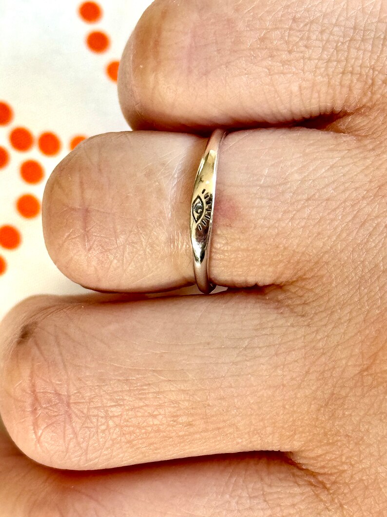 May include: A thin silver band ring with an eye design engraved on the top. The ring is worn on a finger.