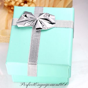 May include: A light blue gift box with a silver ribbon and bow. The box is tied with a silver ribbon and has a silver bow on top. The box is sitting on a wooden surface.