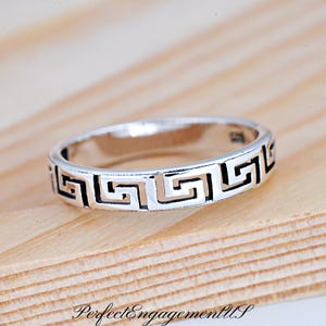 May include: A silver band ring with a Greek key design. The ring is on a wooden surface.