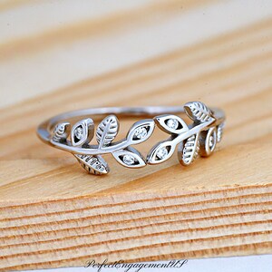 May include: A silver ring with a leaf design. The ring features multiple leaves with small, round, clear gemstones. The ring is set against a light brown wooden surface. The text "PerfectEngagementUS" is at the bottom.