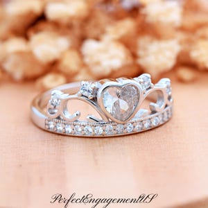 May include: A silver engagement ring with a heart-shaped center stone and a crown design. The ring is adorned with small, clear gemstones and the text "PerfectEngagementUS". The ring is set against a blurred background.