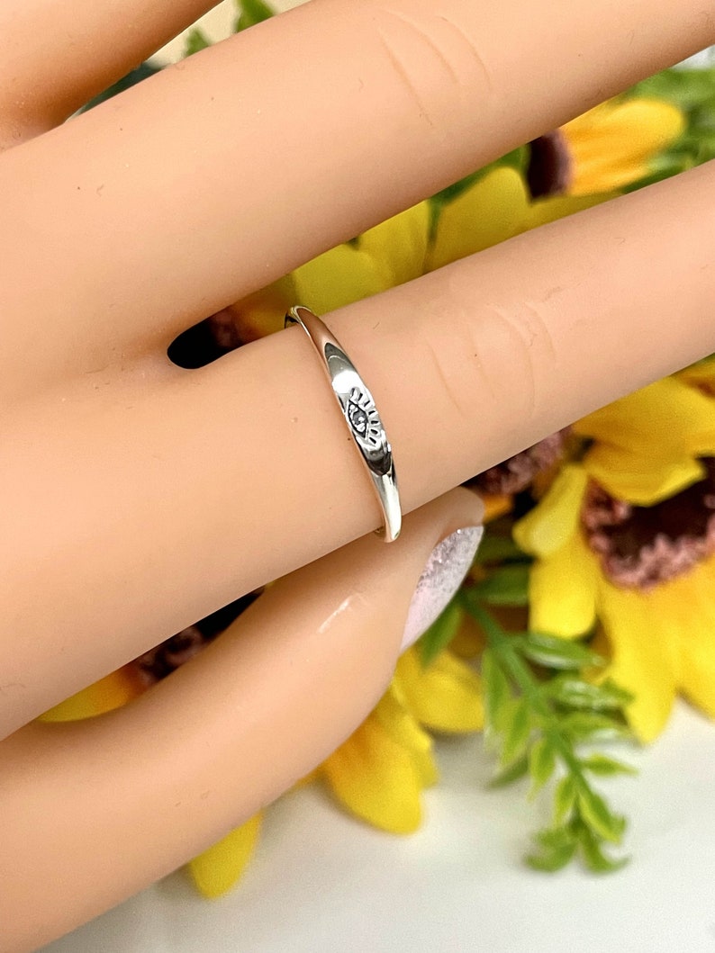 May include: A silver ring with an eye design. The ring is on a finger and the background is blurred.
