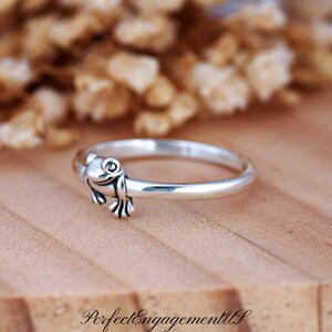 May include: A silver ring featuring a detailed frog design. The frog is positioned on the ring's band, with its head and front legs visible. The ring is set against a wooden surface with a blurred background.
