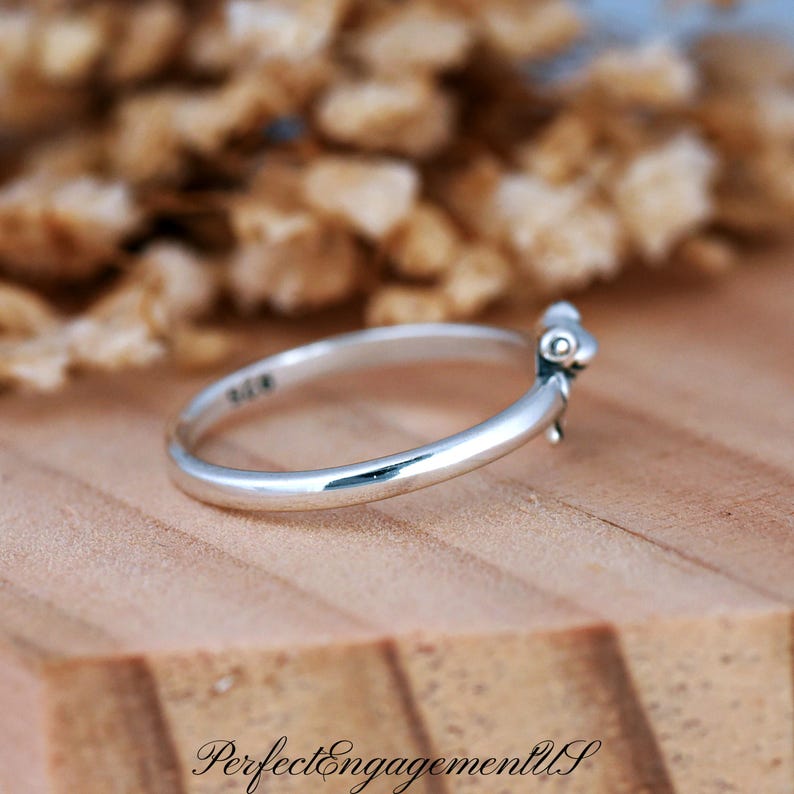 May include: A silver ring with a small, decorative element. The ring is set against a wooden surface with the text "PerfectEngagementUS" at the bottom. The ring's band is smooth and polished.
