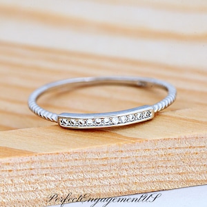 May include: A silver ring with a row of small, clear stones set in a rectangular bar. The band has a twisted rope design. The ring is on a wooden surface. The text "PerfectEngagementUS" is at the bottom.