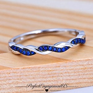 May include: A silver-toned ring featuring a twisted band design. One strand of the twist is polished metal, while the other is set with a continuous row of small, round, vibrant blue stones. The ring is displayed on a light wooden surface.