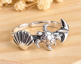 May include: A silver-toned open ring featuring a nautical design with a sea turtle in the center, flanked by a seashell on one side and a ship anchor on the other. The metal has a polished finish with darkened recessed details to highlight the shapes.