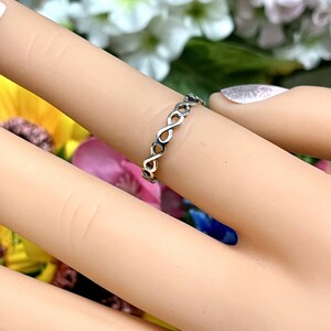 May include: A silver infinity ring with a delicate, braided design. The ring is worn on a finger with a light pink nail polish.