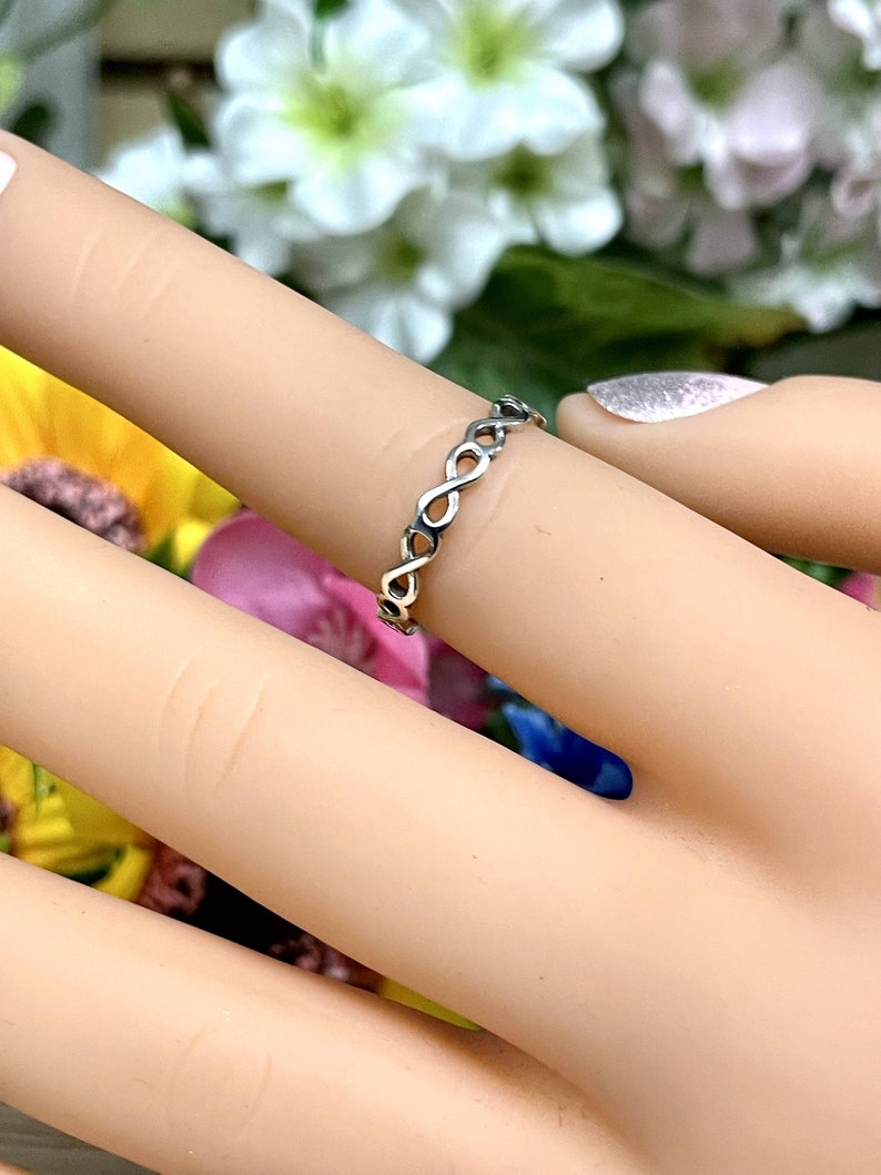 May include: A silver infinity ring with a delicate, braided design. The ring is worn on a finger with a light pink nail polish.