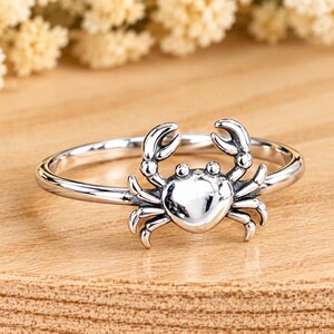May include: A polished silver-toned ring featuring a small, detailed crab ornament centered on the thin band. The crab design includes two raised claws and multiple legs extending from a smooth, rounded shell, creating a cohesive metallic piece.