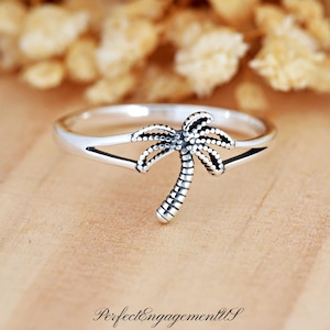 May include: A silver ring featuring a palm tree design. The ring has a split band and a detailed palm tree with a textured trunk and fronds. The ring is set against a light brown surface with blurred background elements.
