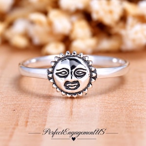 May include: A silver ring featuring a central sun motif with a detailed face and radiating rays. The metal has a polished finish with darkened recessed areas to highlight the facial features of the sun design on the band.