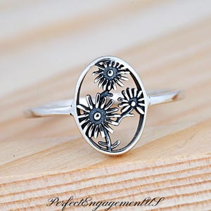May include: A silver ring with an oval-shaped design featuring three stylized flowers with black centers.