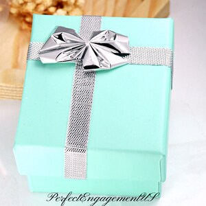 May include: A light blue gift box with a silver ribbon and bow. The box is tied with a silver ribbon and has a silver bow on top. The box is sitting on a wooden surface.