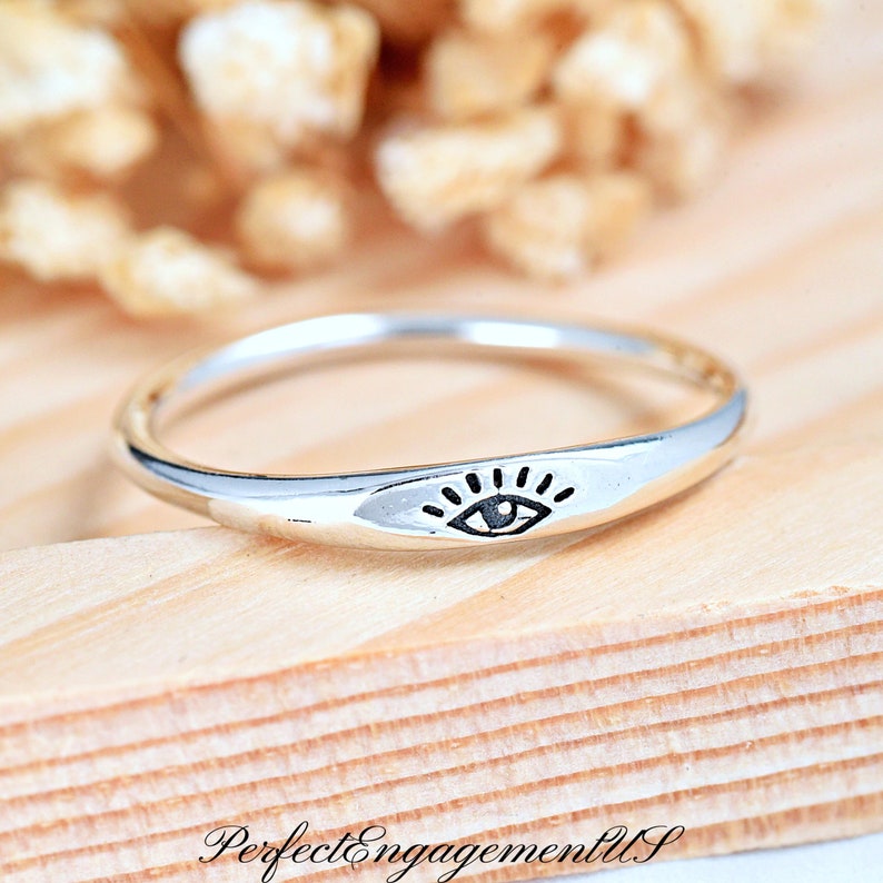 May include: A silver ring with a simple, minimalist design featuring a black eye symbol. The ring is on a wooden surface.