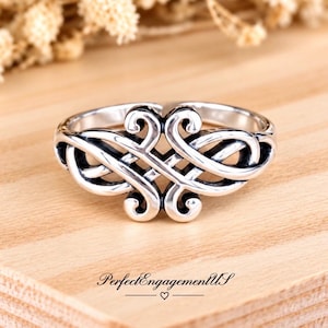May include: A silver ring featuring an intricate, symmetrical openwork design with swirling, interconnected loops and polished metal surfaces. The ring is centered on a light wood surface with the text PerfectEngagementUS printed below it.