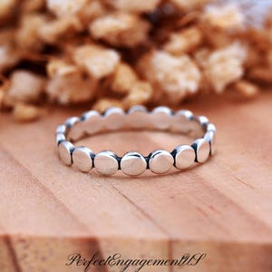 May include: A silver ring with a series of small, round, raised dots around the band. The ring is set against a blurred background of dried flowers and a wooden surface. The text "PerfectEngagementUS" is visible at the bottom.