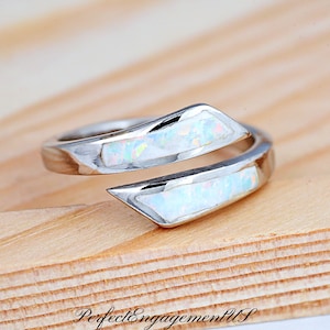 May include: A silver ring with a wrap-around design. The ring features two sections of white opal stone with iridescent flecks. The ring is set on a wooden surface.