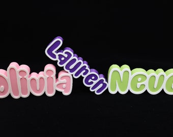 May include: Three 3D printed names, Olivia, Lauren, and Neve, are displayed on a black background. The names are in different colors: Olivia is pink, Lauren is purple, and Neve is green.