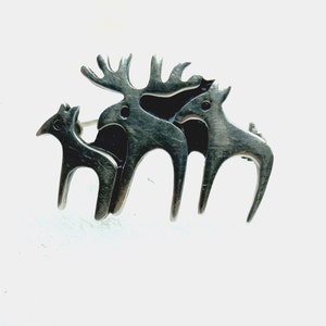 May include: A silver brooch featuring three stylized reindeer, one larger than the others. The reindeer are facing the right side of the image.
