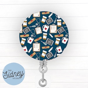May include: A round, navy blue badge reel with a repeating pattern of medical supplies. The pattern includes bandages, stethoscopes, pill bottles, clipboards, mugs with hearts, and syringes. The badge reel has a clear plastic strap and a metal clip. The text "The Sidney Studio" is on a light blue circle.
