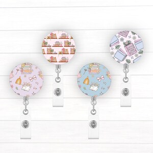 May include: Four retractable badge holders with different book-themed designs. The designs feature stacks of books, flowers, and bows in pink, blue, and white on a light blue or pink background.