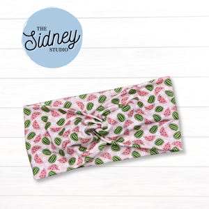 May include: A pink headband with a repeating pattern of green watermelons and white flowers. The headband has a twisted knot design in the center. The text "The Sidney Studio" is in a light blue circle in the upper left corner.
