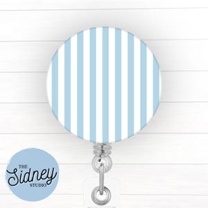 May include: A circular badge reel featuring a pattern of vertical light blue and white stripes. The device includes a clear plastic strap with a metal snap button and a retractable cord mechanism, designed for holding identification cards or keys.
