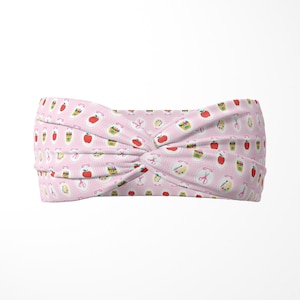 May include: A pink headband with a knotted design. The headband is patterned with various illustrations, including apples, scissors, and drinks. The background features vertical stripes. This accessory is designed for comfort and style.