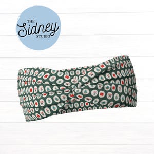 May include: A green headband with a repeating pattern of white circles containing red gift boxes and green Christmas trees. The headband has a knotted design and the text "The Sidney Studio" in a light blue circle.
