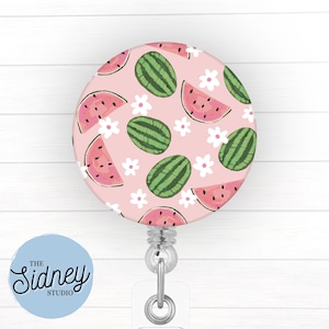 May include: A round retractable badge reel with a pink background featuring a pattern of green watermelons, pink watermelon slices, and white flowers. The badge reel has a clear plastic strap and a metal clip. The text "The Sidney Studio" is on a light blue circle.