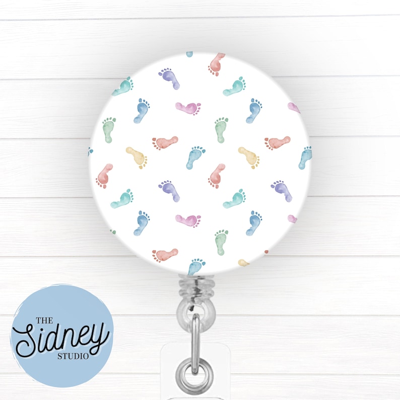 Puede incluir: Un carrete de insignia retr&aacute;ctil blanco con un patr&oacute;n de huellas de beb&eacute; en colores pastel. El carrete tiene un clip plateado y una correa de pl&aacute;stico transparente. El texto "The Sidney Studio" est&aacute; en un c&iacute;rculo azul claro.